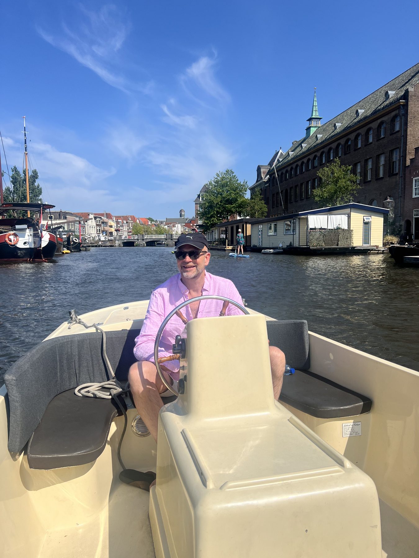 Alex at the wheel, pink shirt, sunglasses, and the quiet confidence of a man who has steered exactly one (1) canal boat.